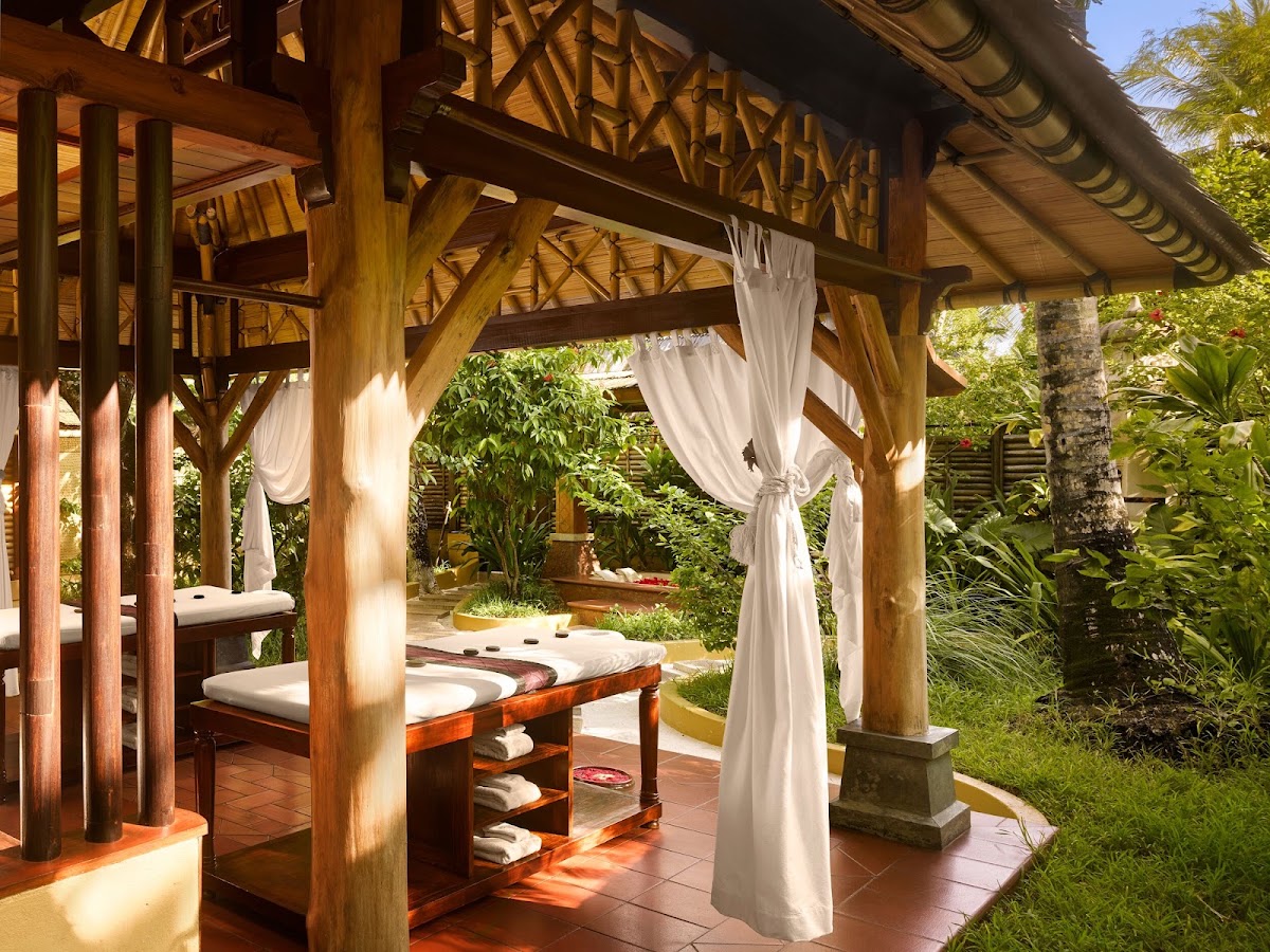 Wooden pavilion with white curtains draped from beams, massage tables with rolled towels, and lush tropical plants surrounding the spa area.