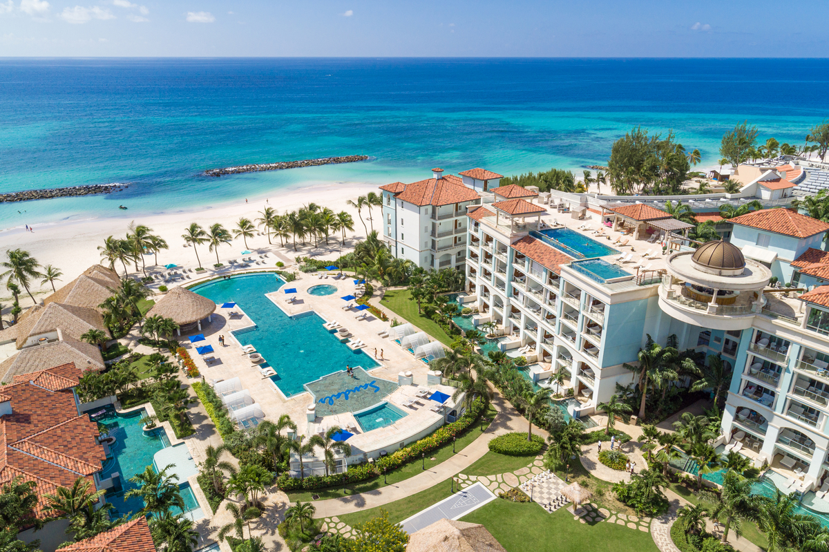 Aerial view of Sandals Royal Barbados with multiple pools, white-roofed buildings, palm trees, and turquoise Caribbean waters beyond the white sand beach.