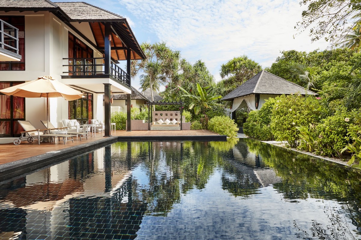 Infinity pool reflecting white colonial-style villas with dark trim, loungers and umbrella on wooden deck, lush tropical gardens and palm trees surrounding.