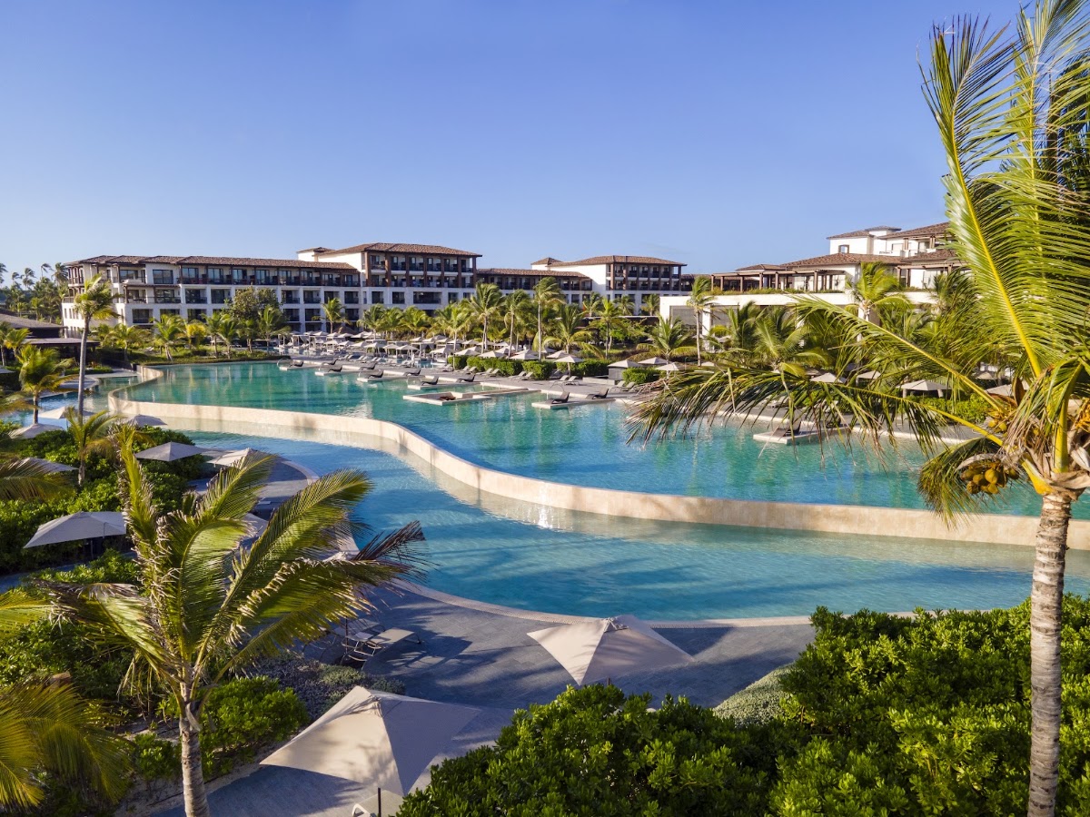Large lagoon-style pool with turquoise water and sand beach surrounded by palm trees, lounge chairs, and multi-story resort buildings under clear sky.