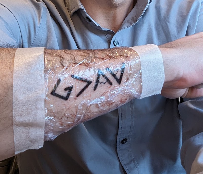 G>∧∨ symbolen tattoo op onderarm, vers gezet met folie.