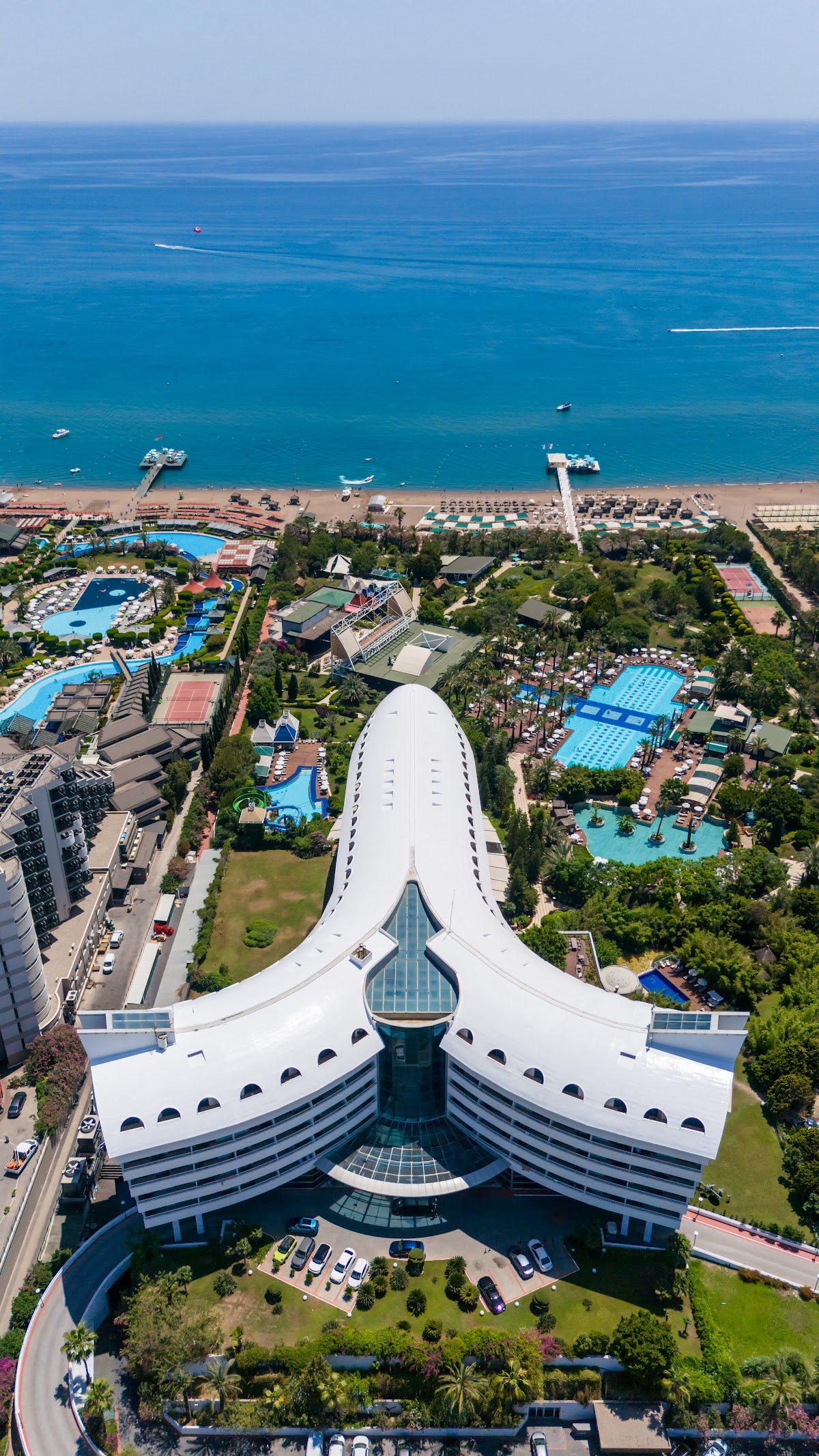 Aerial view of Concorde De Luxe Resort's distinctive white building with curved wings overlooking multiple pools, beach, and turquoise Mediterranean Sea with boats.