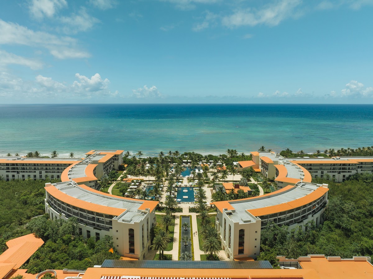 Aerial view of UNICO 20°87° Hotel Riviera Maya with curved orange-roofed buildings framing multiple pools, surrounded by palm trees and tropical vegetation, overlooking turquoise Caribbean waters.