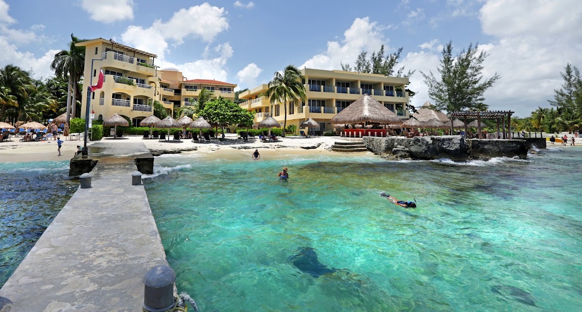 Playa Azul Cozumel Hotel's yellow buildings overlook a sandy beach and clear turquoise water with swimmers and a stone pier in foreground.