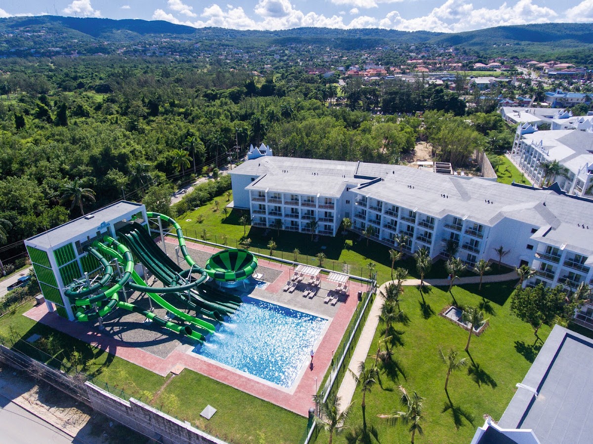 Aerial view of white resort buildings with a pool featuring bright green water slides, surrounded by manicured lawns and forested mountains beyond.