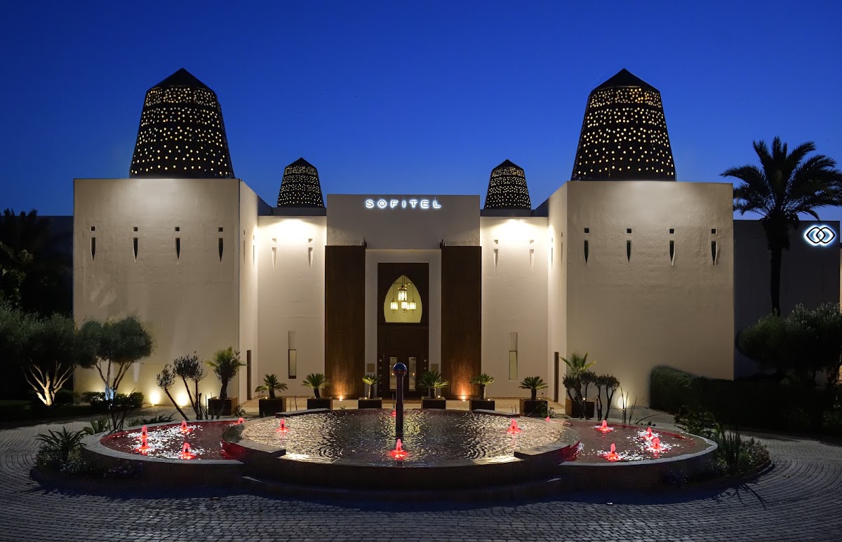 Sofitel Agadir Royal Bay Resort's illuminated entrance at dusk features cream-colored towers with perforated tops, a central archway, and a circular courtyard pond with red candles and flowering plants.