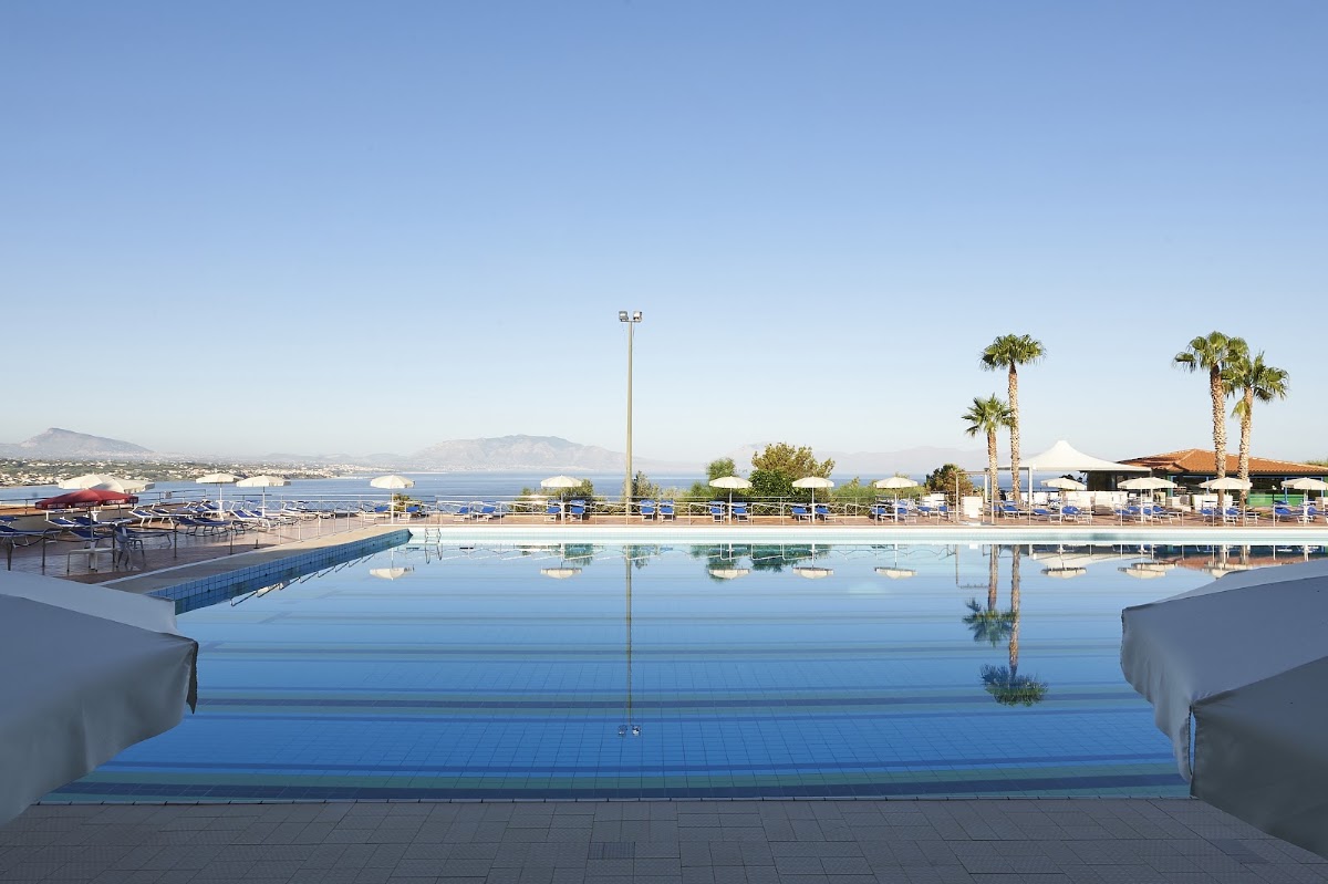 Large blue swimming pool overlooking the Mediterranean Sea with palm trees, loungers, and white umbrellas under clear sky.