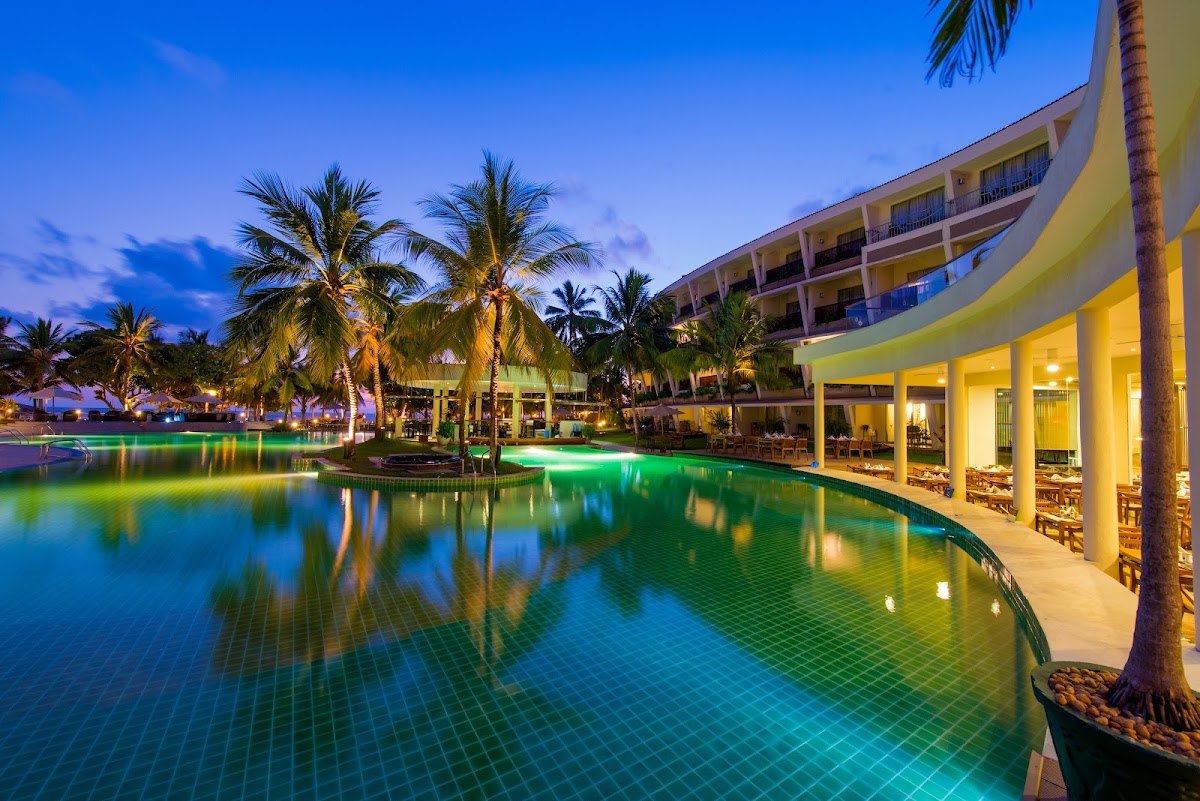 Illuminated pool with green lighting at dusk, curved yellow resort building with lit columns, palm trees, and covered seating areas at The Eden Beruwala.