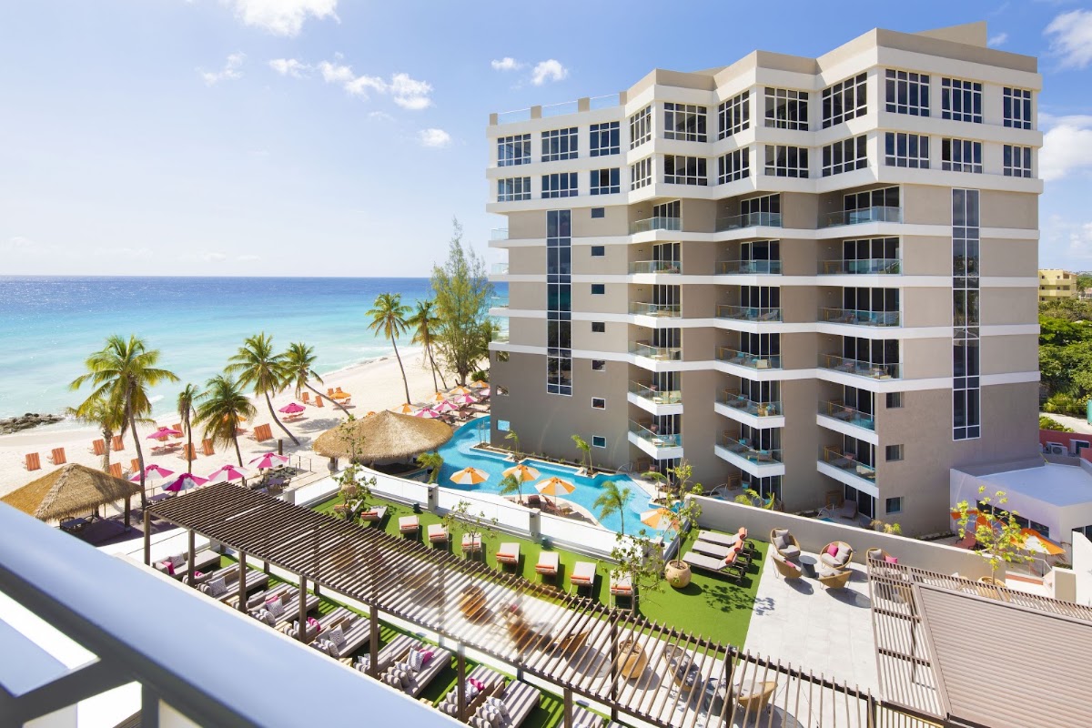 Multi-story beachfront resort with pool, lounge chairs, and palm-lined sand leading to turquoise Caribbean waters.