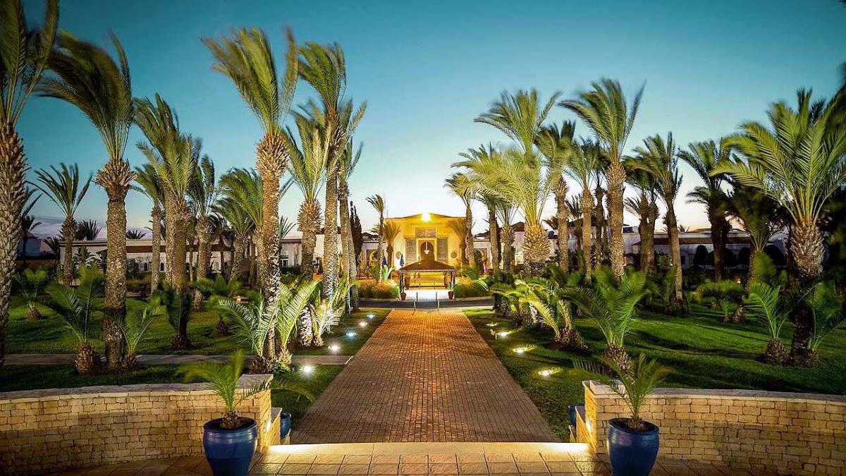 Illuminated brick pathway lined with palm trees leads to a glowing yellow building at dusk at ROBINSON Agadir.