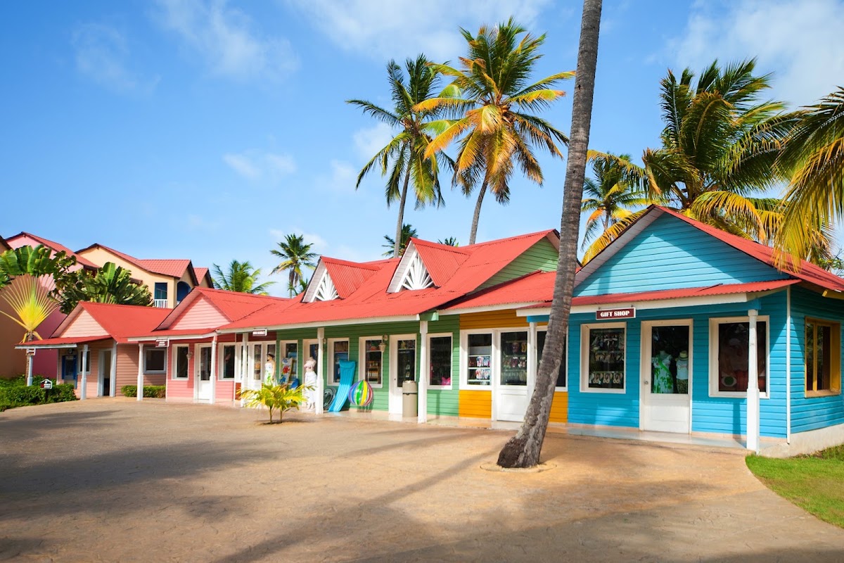 Colorful beachfront shops with red roofs and pastel walls line a sandy courtyard dotted with palm trees at Punta Cana Princess.