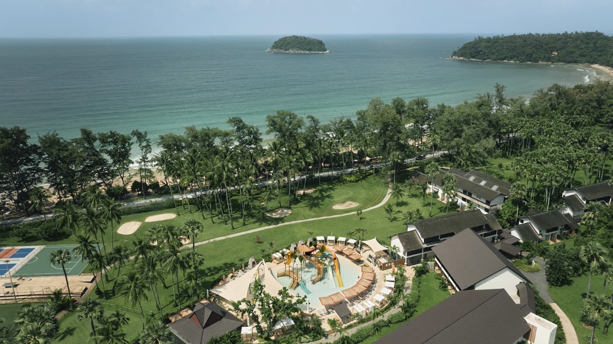 Young child slides down orange water slide while adults relax in shallow pool surrounded by palm trees and tan umbrellas.