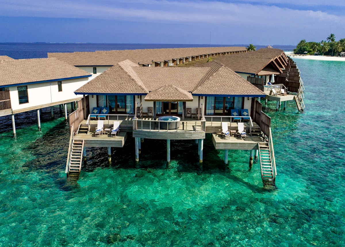 Overwater bungalows with wooden decks and thatched roofs at Reethi Faru Resort, surrounded by turquoise lagoon and distant island.