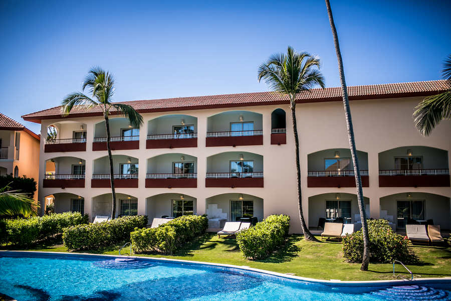 Majestic Colonial Punta Cana's cream-colored building with red-trimmed balconies overlooks a blue pool, manicured lawn, and palm trees under clear sky.
