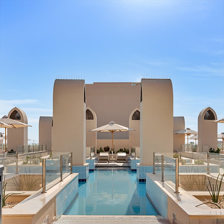 Rectangular pool with cream umbrellas and loungers flanked by tan architectural structures with arched windows under clear blue sky.
