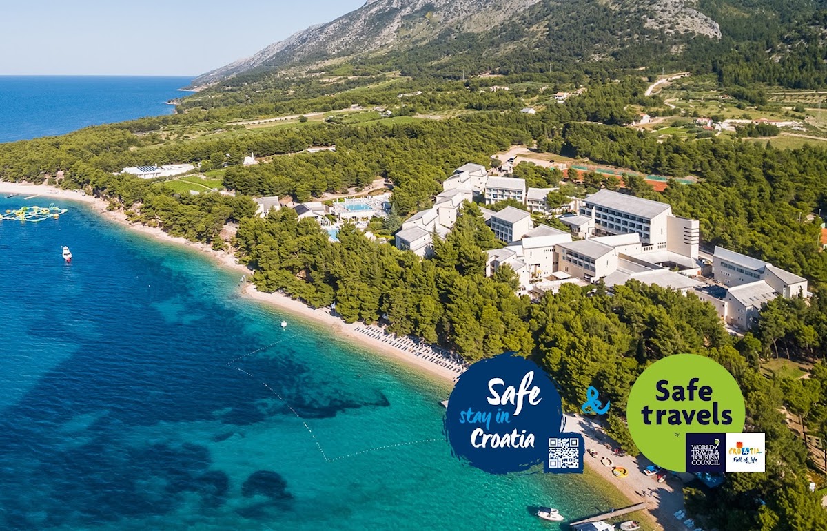 Aerial view of Bretanide Sport & Wellness Resort nestled between forested hillside and turquoise Adriatic Sea with pebbly beach below.