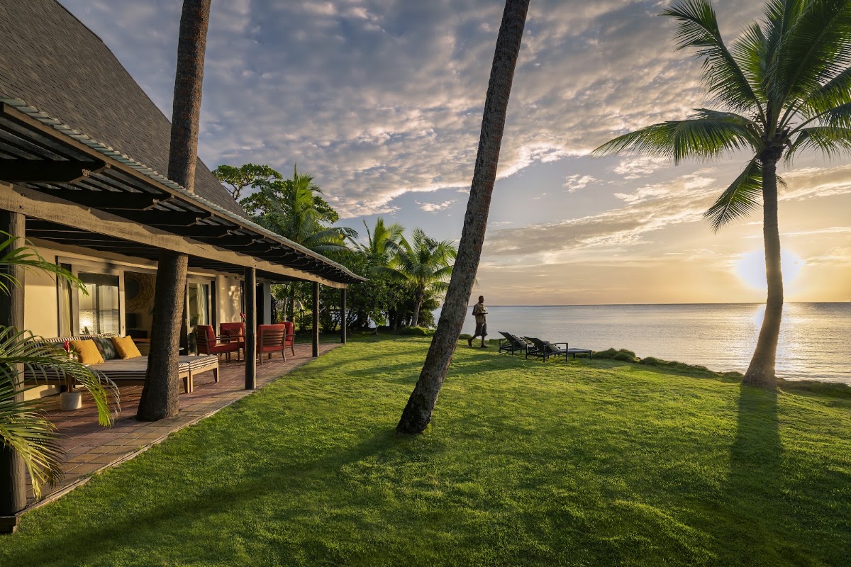 Thatched bungalow with wooden deck overlooks green lawn and palm trees toward sunset ocean at Shangri-La Yanuca Island.