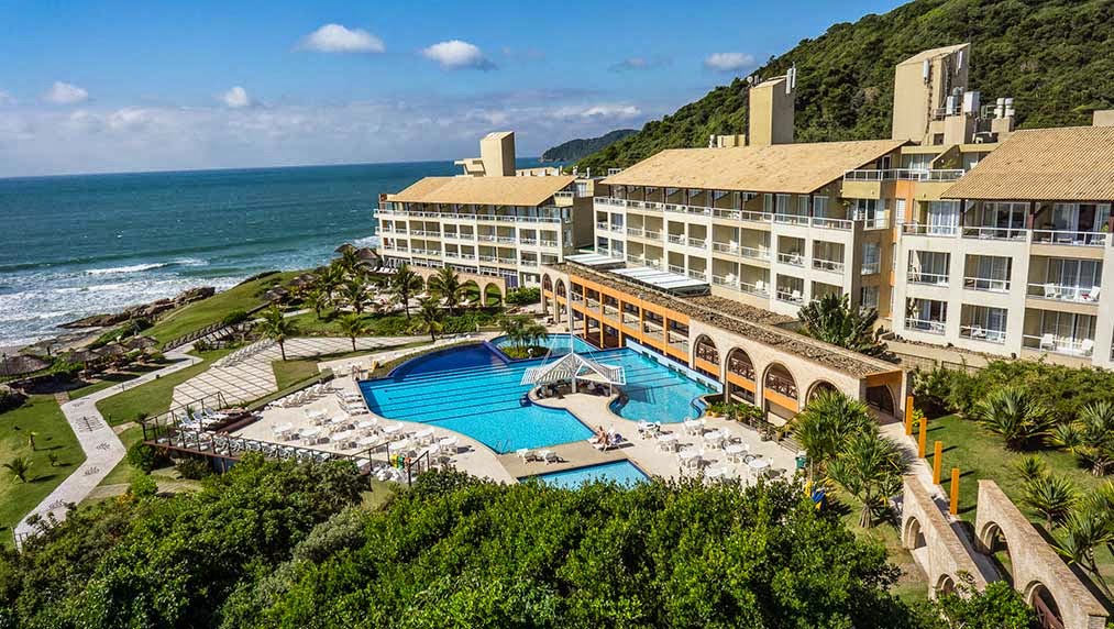 Costao do Santinho Resort sits on a hillside overlooking the ocean, with a large blue pool, loungers, and multi-story beige buildings surrounded by lush green vegetation.