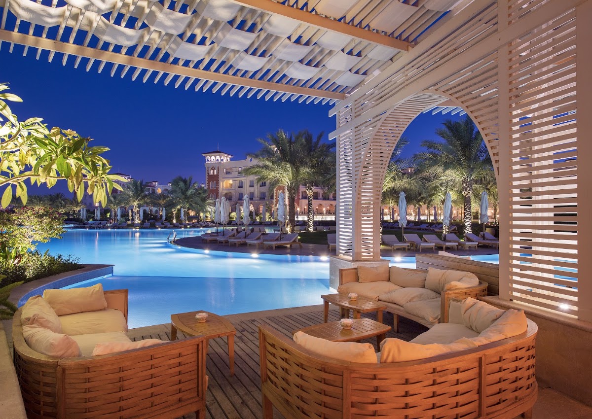 Twilight view from a covered lounge at Baron Palace Sahl Hasheesh showing illuminated pool, palm trees, and palace-style building beyond.