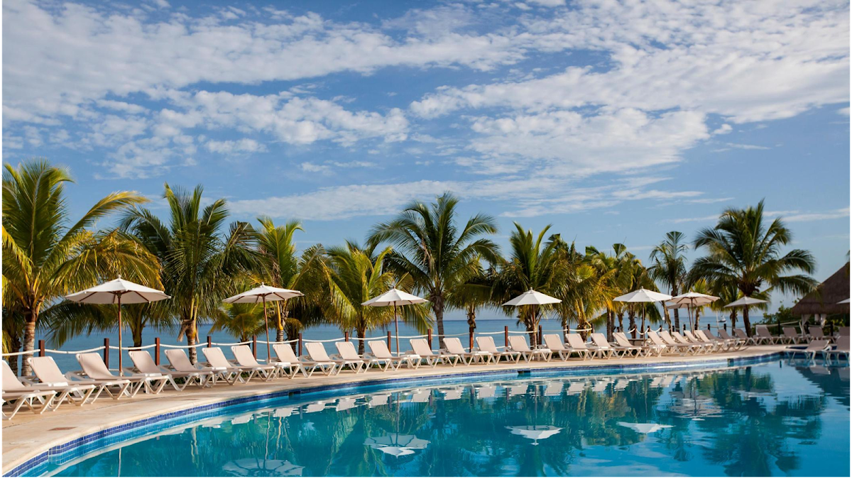 Beachfront pool at Occidental Cozumel lined with white loungers and umbrellas, palm trees, and calm turquoise water beyond.