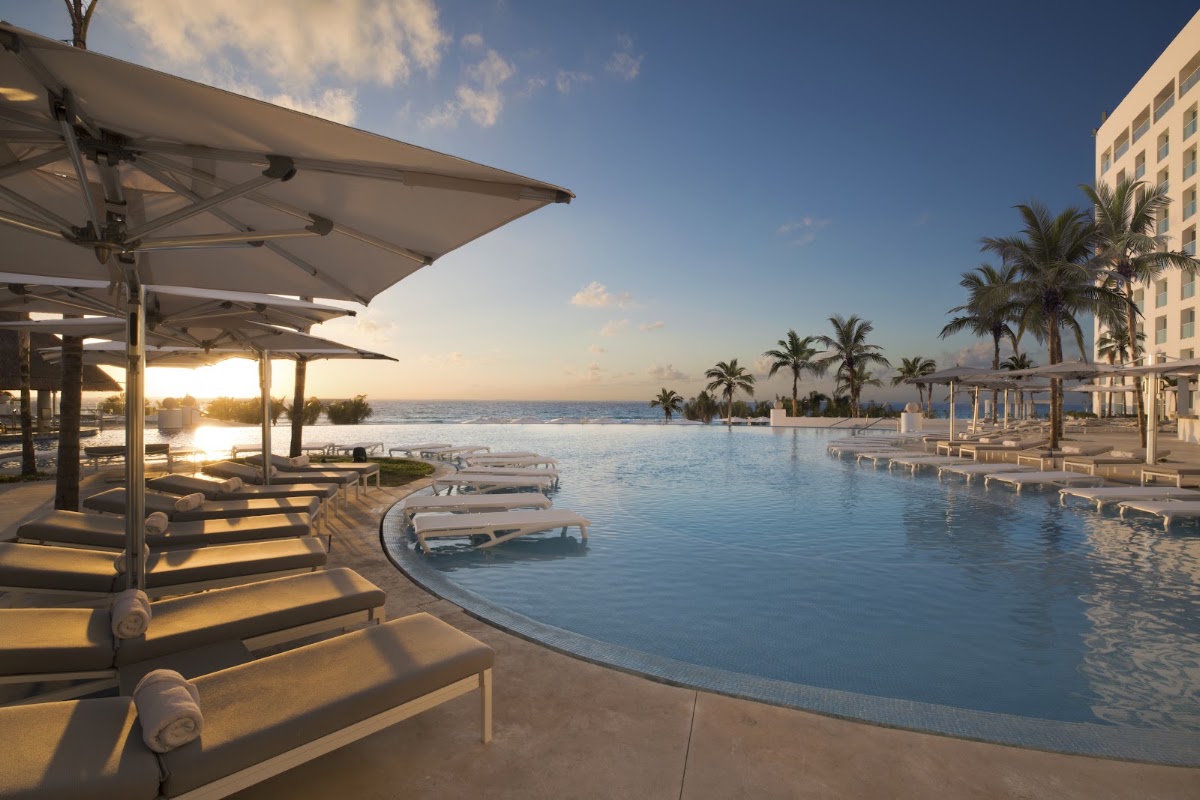Shaded loungers and umbrellas line the pool deck at Le Blanc Spa Resort Cancun, overlooking the ocean at sunset with palm trees and the main building visible.