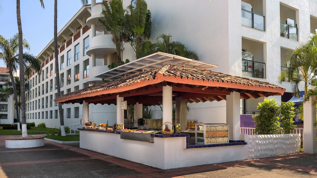 Outdoor dining pavilion with red clay tile roof and white columns at Meliá Puerto Vallarta, with modern resort building and palm trees behind it.