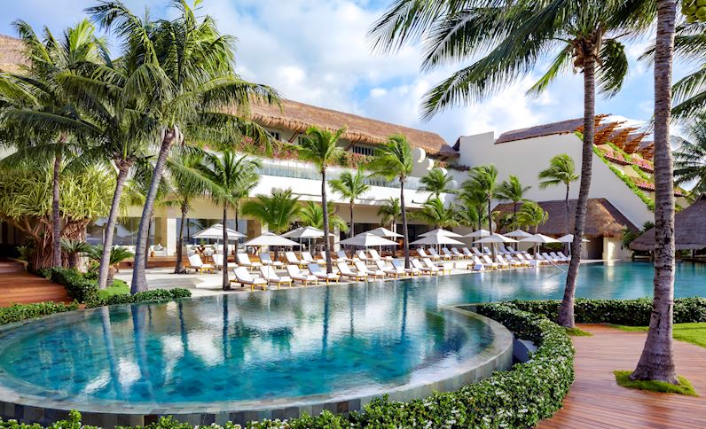 Curved pool with turquoise water surrounded by palm trees, white lounge chairs, and thatched-roof buildings at Grand Velas Riviera Maya.
