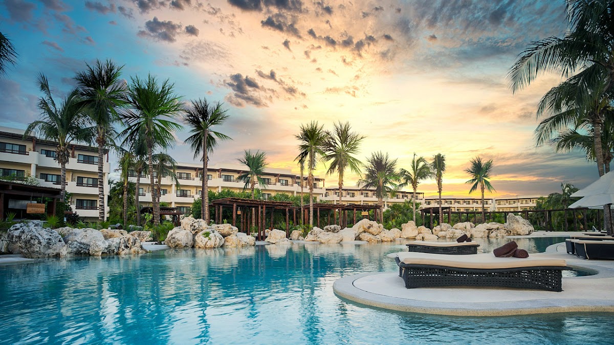 Sunset over turquoise lagoon at Secrets Maroma Beach Riviera Cancun with lounge chairs, palm trees, and whitewashed resort buildings.