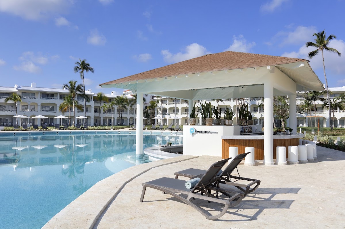 Two lounge chairs on the pool deck facing a modern white pavilion bar with a wooden counter and potted cacti, palm trees visible beyond white resort buildings.