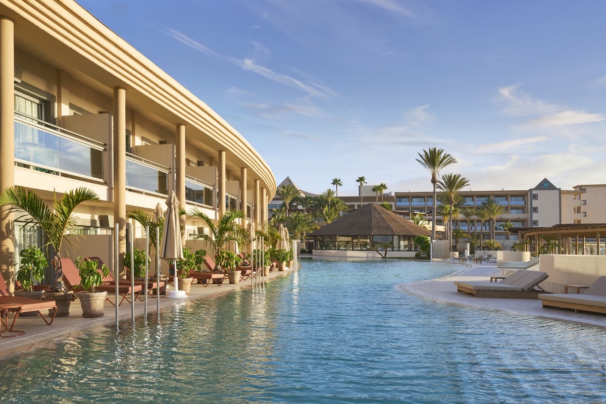 Resort pool with curved tan building, thatched-roof bar, palm trees, lounge chairs, and multi-story accommodations under blue sky.