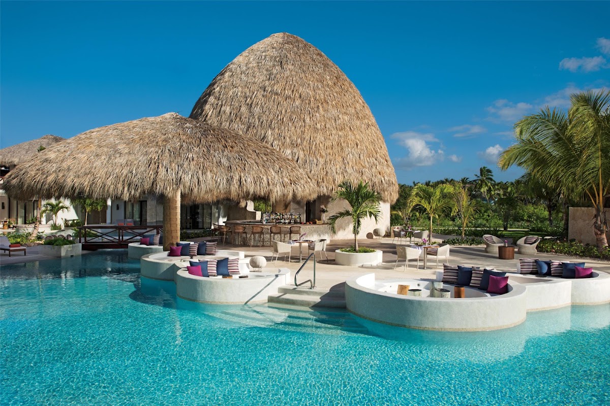 Beachfront bar with a thatched palapa roof, white columns, and wooden counter on the sand at Secrets Cap Cana Resort & Spa.