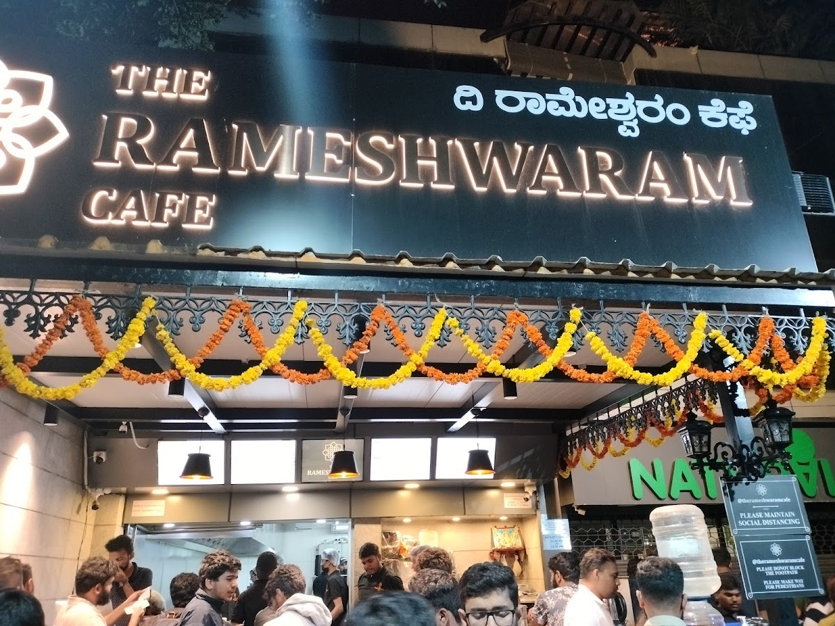 The Rameshwaram Cafe - Indiranagar's vibrant exterior welcomes guests with colorful floral garlands and modern signage, reflecting its lively and inviting character.