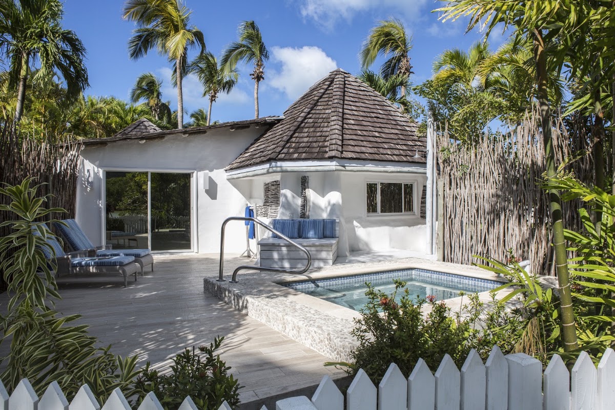 White villa with dark conical roof surrounded by tall palms, featuring a private hot tub on the deck and lounge chairs beside the building.