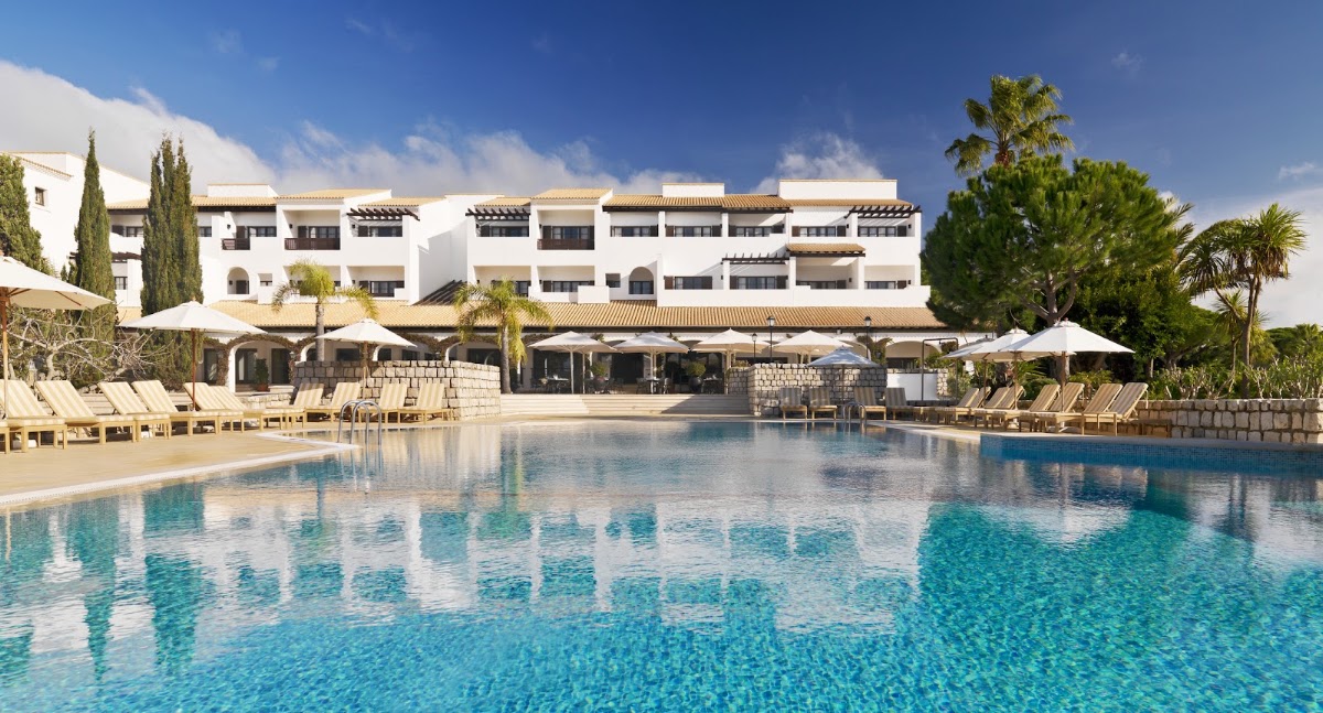 Resort's large swimming pool with clear turquoise water, loungers, white umbrellas, and whitewashed buildings with tiled roofs against blue sky.