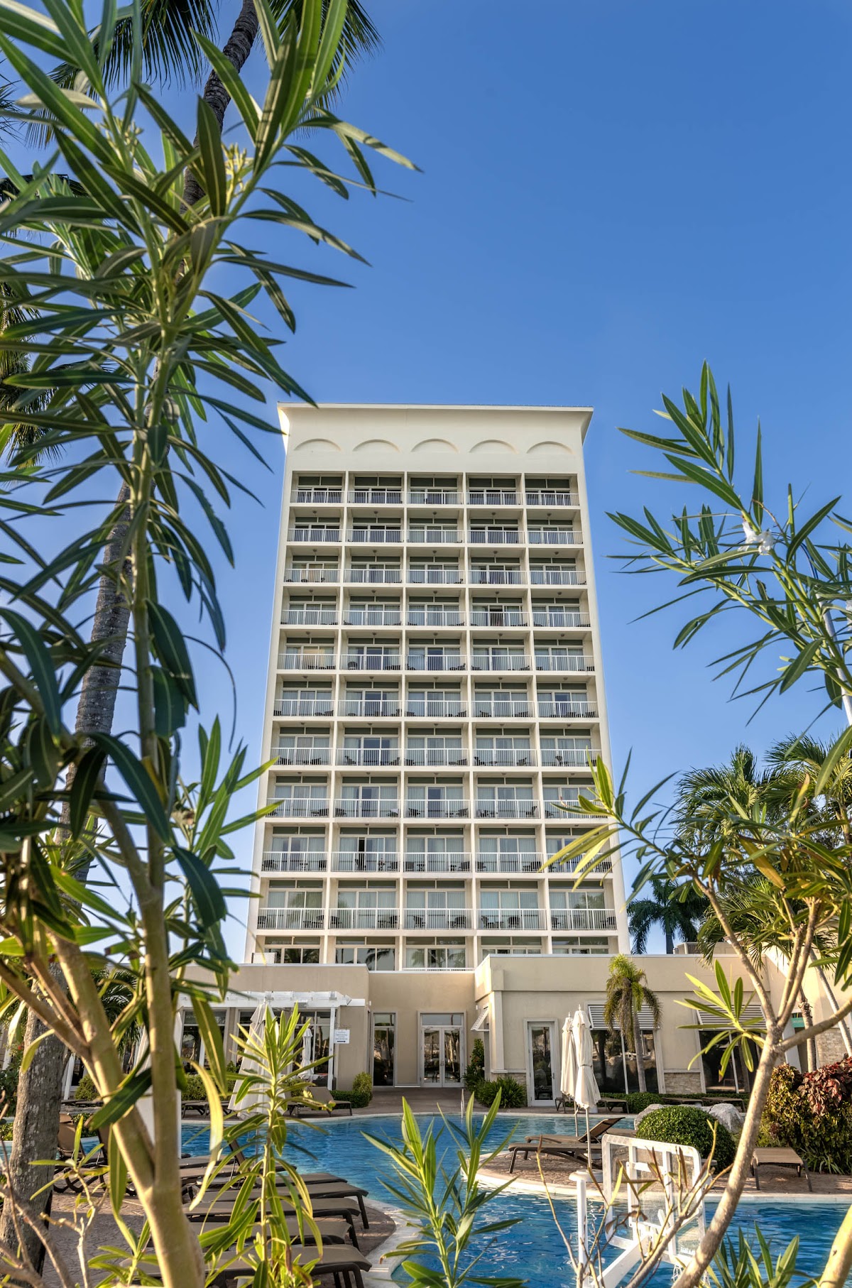 Tall cream-colored hotel tower with balconied rooms overlooks a blue pool surrounded by tropical plants and lounge chairs under clear sky.