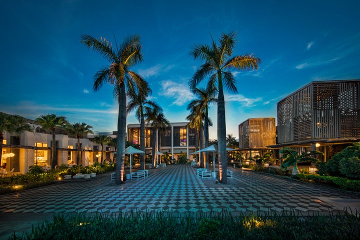 Tall palm trees line a patterned courtyard at Long Beach Mauritius, with illuminated modern bungalows and buildings flanking both sides under a twilight sky.