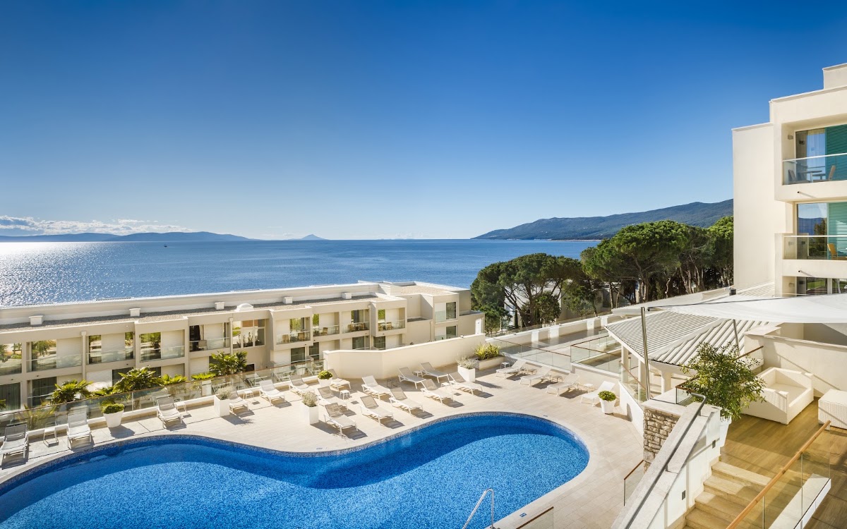 Curved blue pool with lounge chairs on white terrace overlooking Adriatic Sea and mountains at Valamar Bellevue Resort.