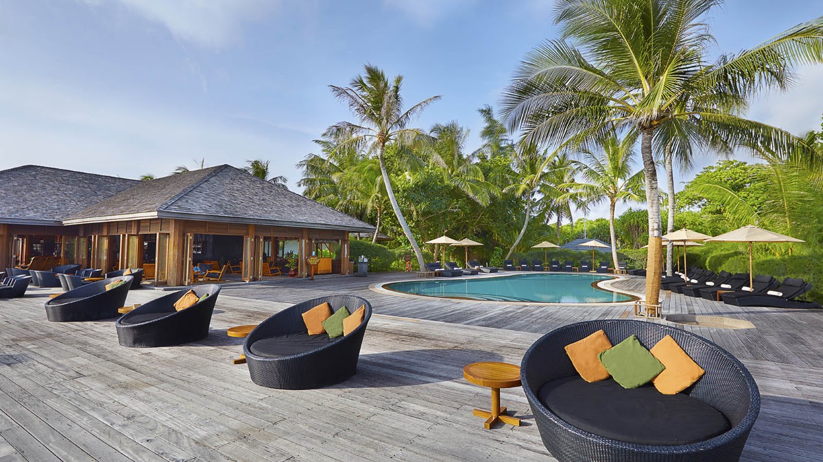 Wooden deck with black round lounge chairs and colorful cushions, resort building and pool surrounded by palm trees under blue sky at Kuredu Island Resort & Spa.