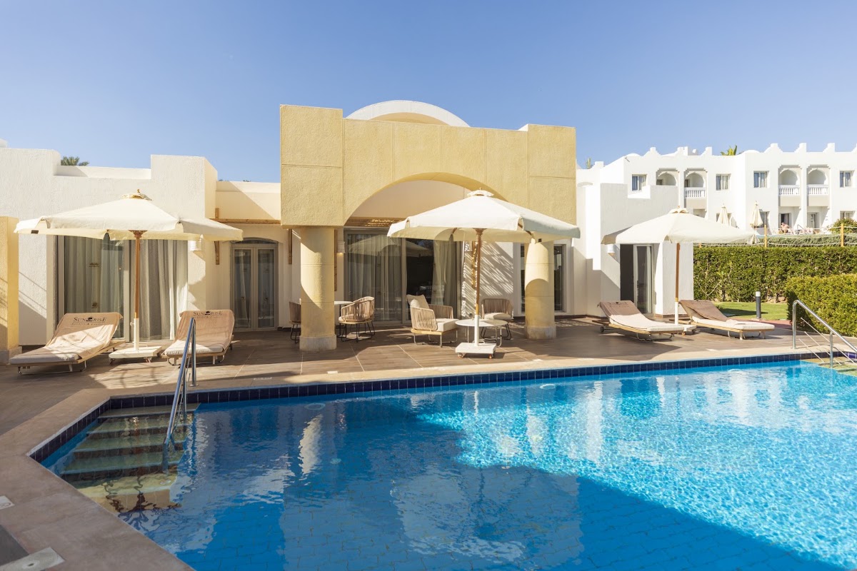 Blue swimming pool with white umbrellas and loungers on the deck, cream and yellow resort buildings with arched architecture behind.