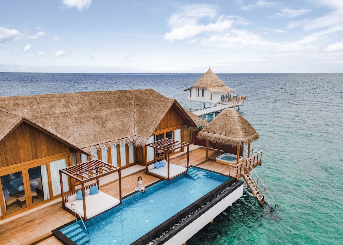Overwater bungalows with thatched roofs and wooden decks at Furaveri Maldives, featuring a shared infinity pool and ocean views.