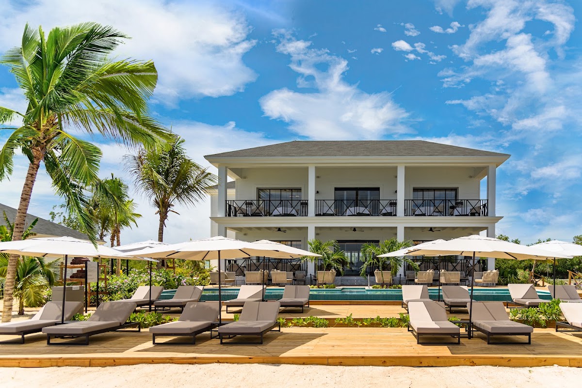 Two-story beachfront resort building with white columns and balconies, lounge chairs and umbrellas on sandy beach, palm trees, and pool visible.