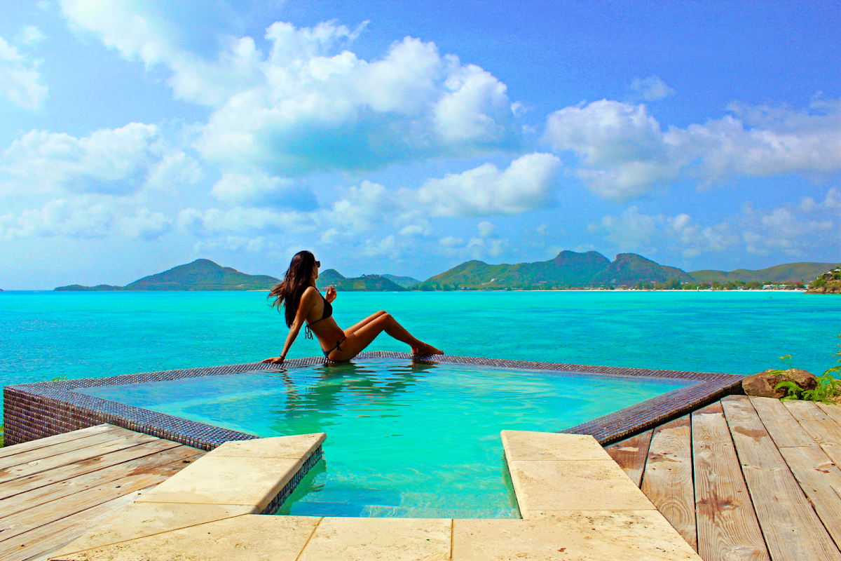 Woman in bikini lounges on mesh platform in shallow turquoise water at Cocobay Resort Antigua, with green volcanic islands visible across the bay.