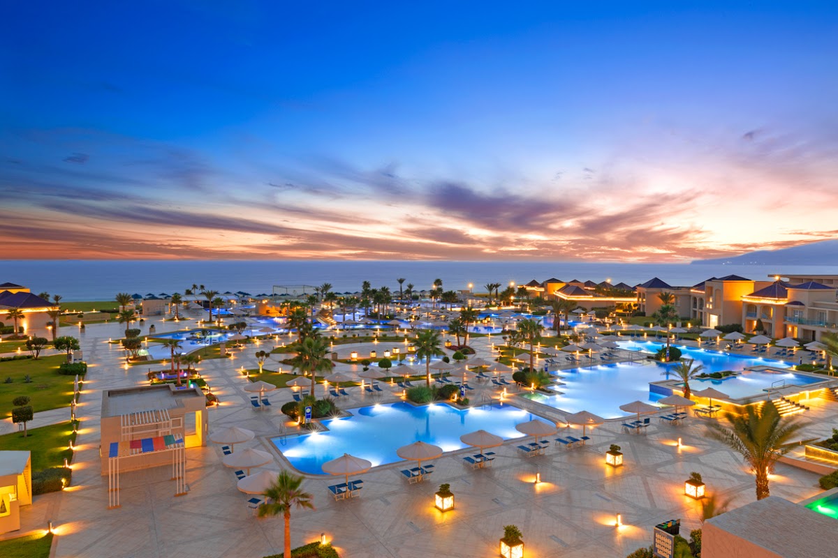 Illuminated pools and palm trees at Pickalbatros White Beach Taghazout at twilight, with sandy grounds and resort buildings under a colorful sunset sky.