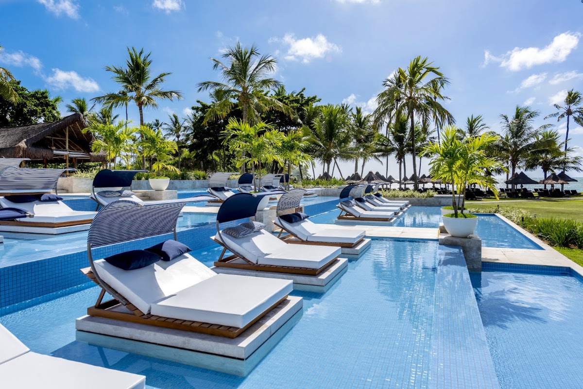 Lounge chairs with canopies line the bright blue pool deck at Tivoli Ecoresort Praia do Forte, with palm trees and beach cabanas visible beyond.