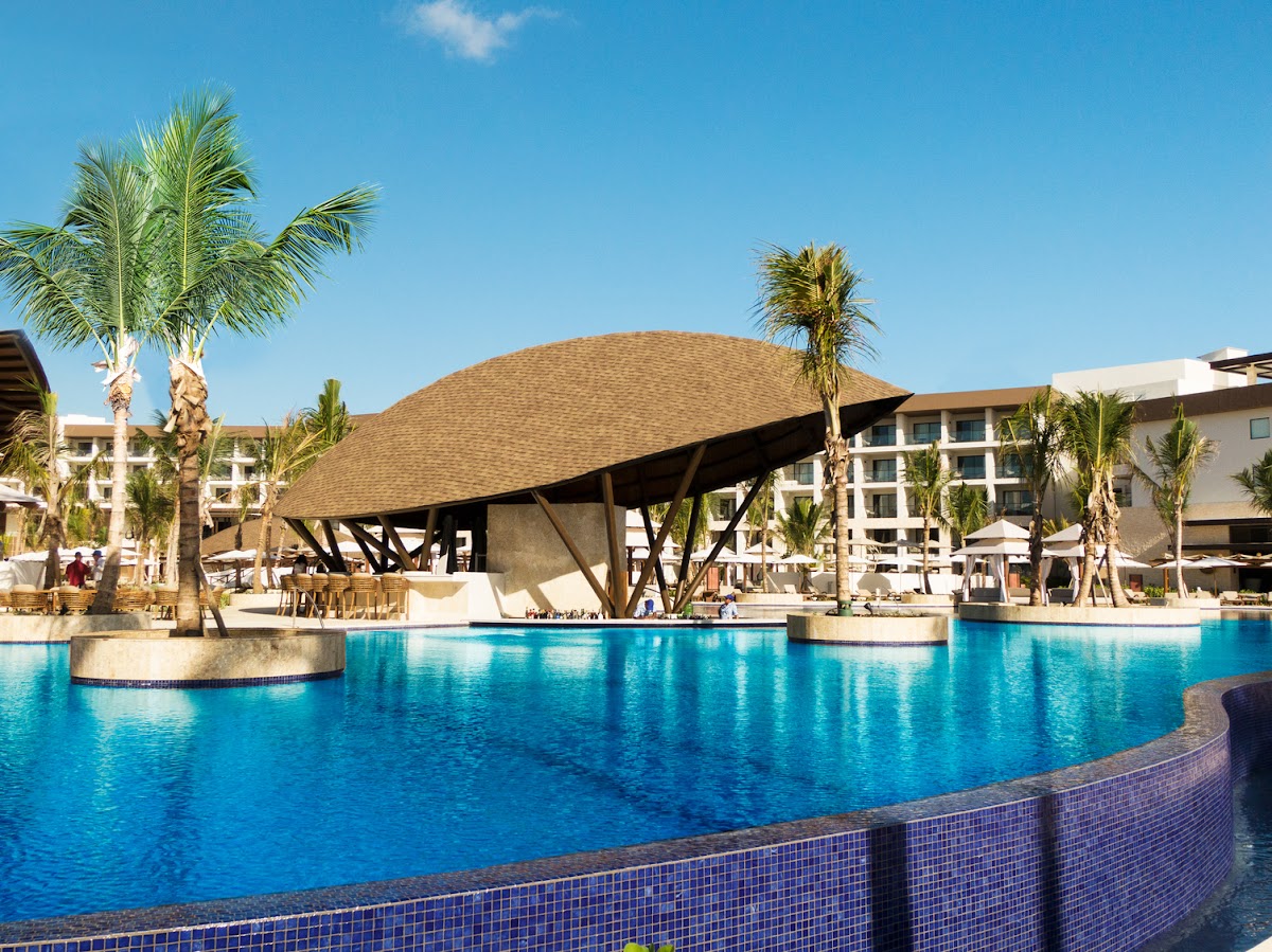 Large curved pool with tiled edge, wooden-roofed palapa bar, palm trees, and white resort buildings under clear blue sky.