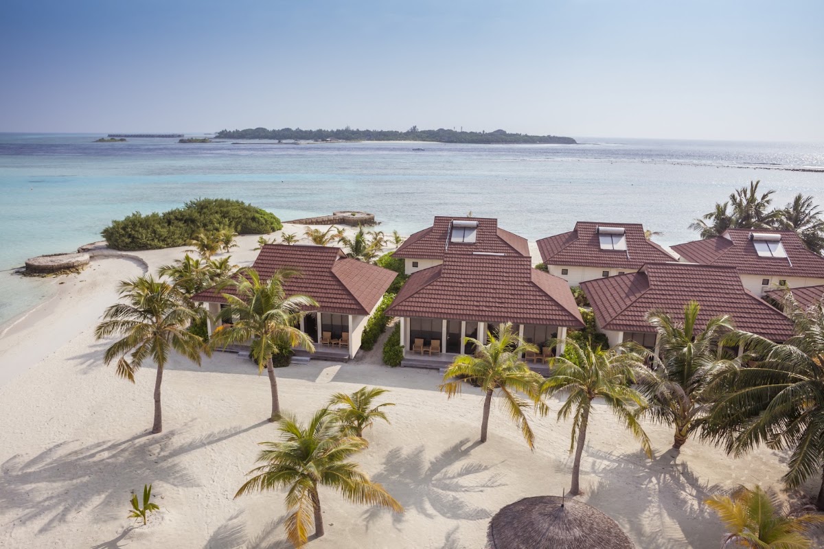 Beachfront villas with red-tiled roofs sit on white sand beside turquoise lagoon water, with palm trees and distant islands visible.