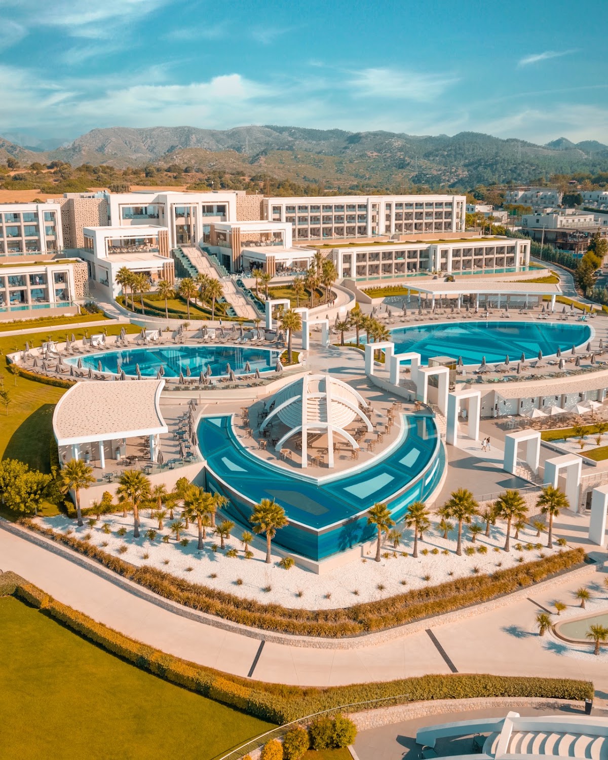 Aerial view of Mayia Exclusive Resort & Spa with white buildings, multiple pools, water slides, loungers, and palm trees against forested mountains.