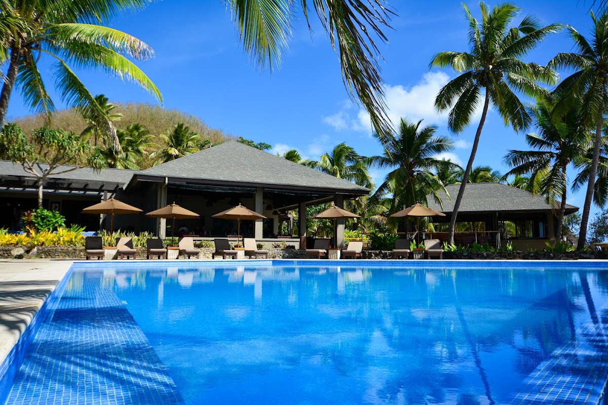 Large blue swimming pool with tan lounge chairs and umbrellas on the deck, dark-roofed pavilion, and palm trees under clear sky at Yasawa Island Resort & Spa.